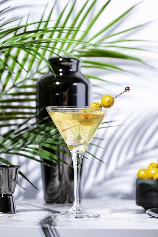 Cocktailbar Lochau: eleganter Martini im Stielglas mit drei Oliven, Shaker und tropischen Palmzweigen im Hintergrund.