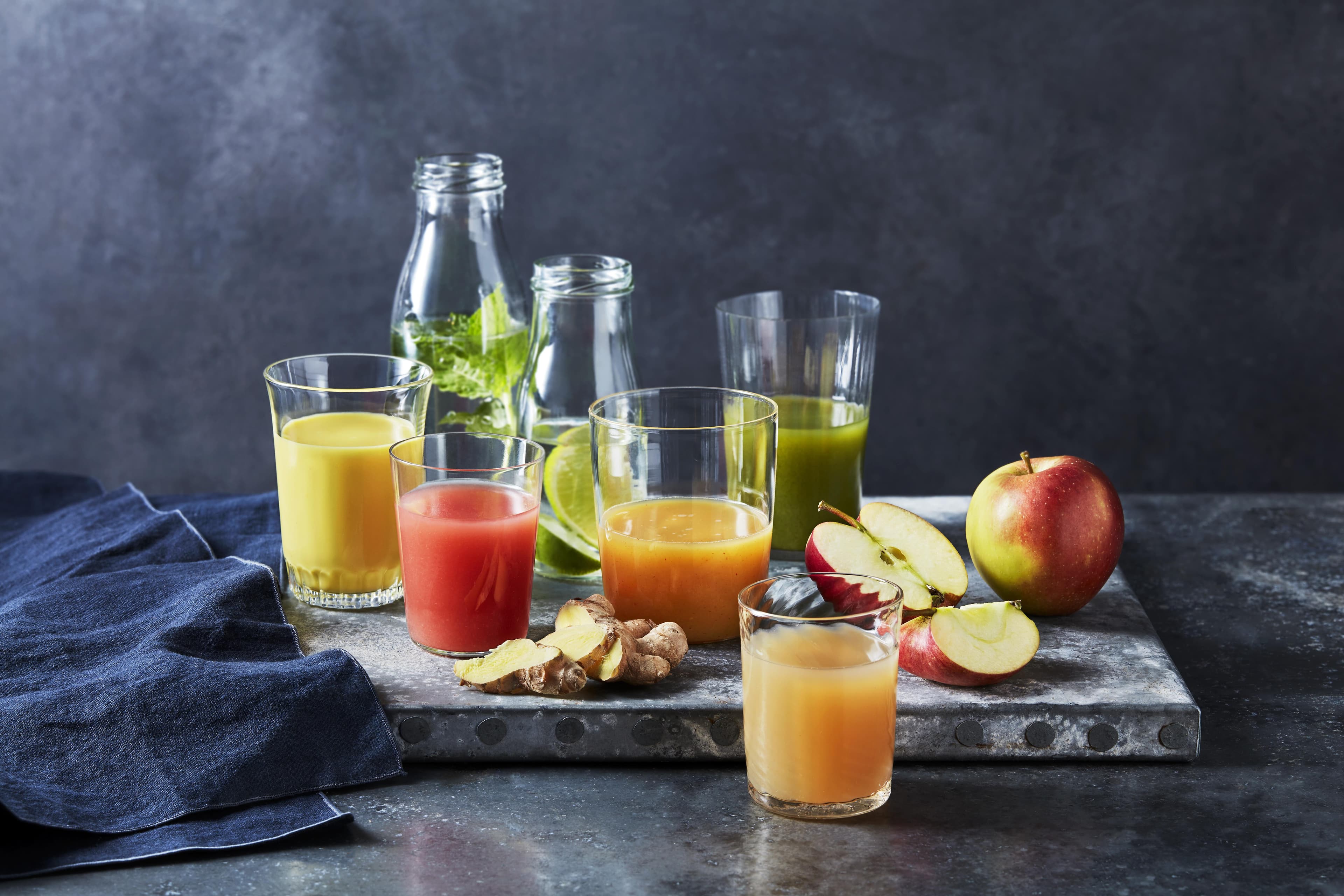 Verschiedene bunte Frucht- und Gemüsesäfte in Gläsern und Flaschen mit frischen Äpfeln und Ingwer auf einem rustikalen Metalltablett.
