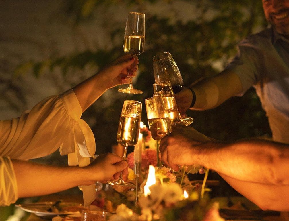 Eine Gruppe stoßt bei einem Abendessen im Freien mit Sektgläsern an, die von Kerzenlicht beleuchtet werden.