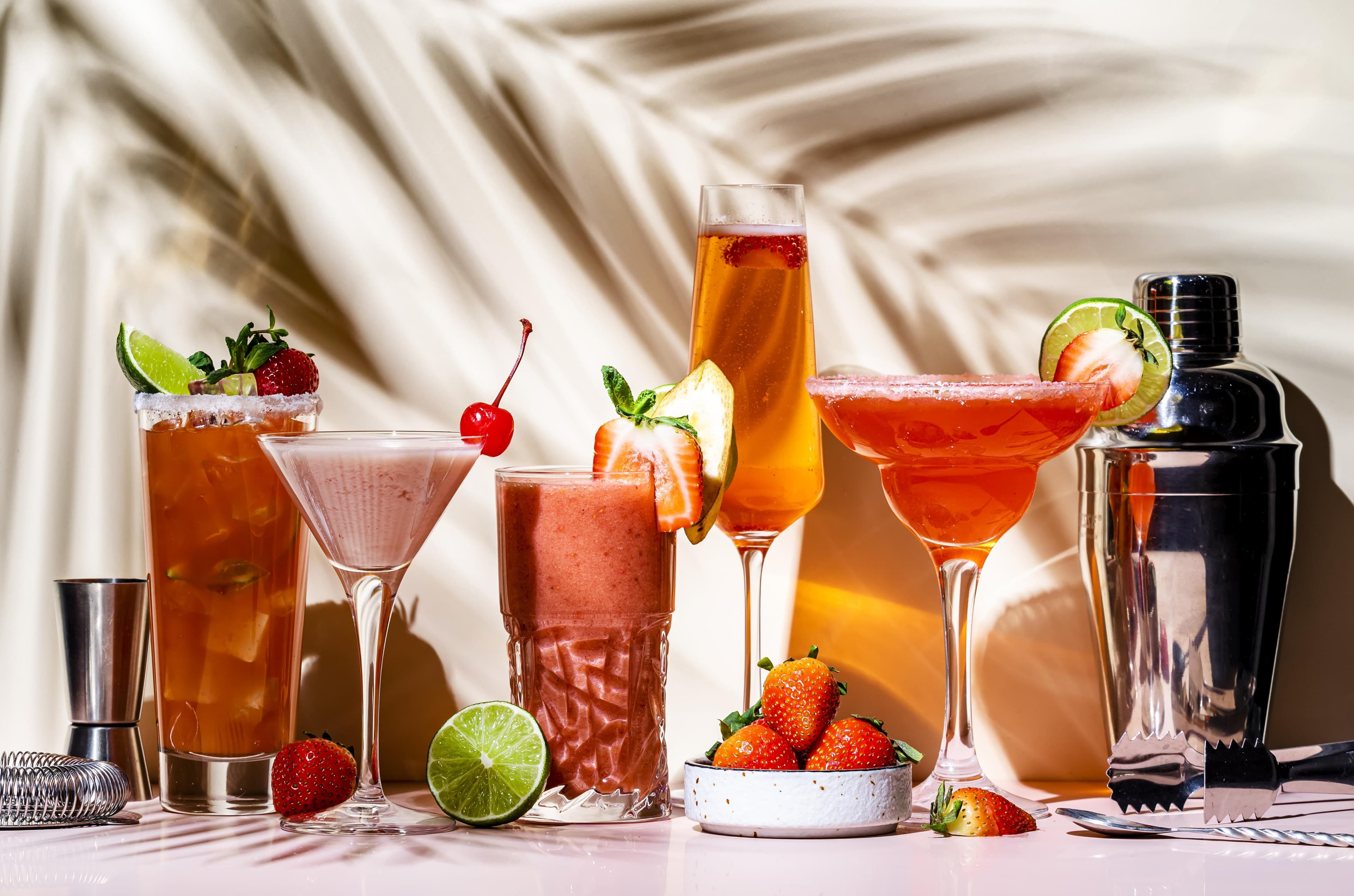 Eine Auswahl bunter Cocktails mit Erdbeeren und Limetten, Bar-Zubehör und tropischen Schattenspielen im Hintergrund.