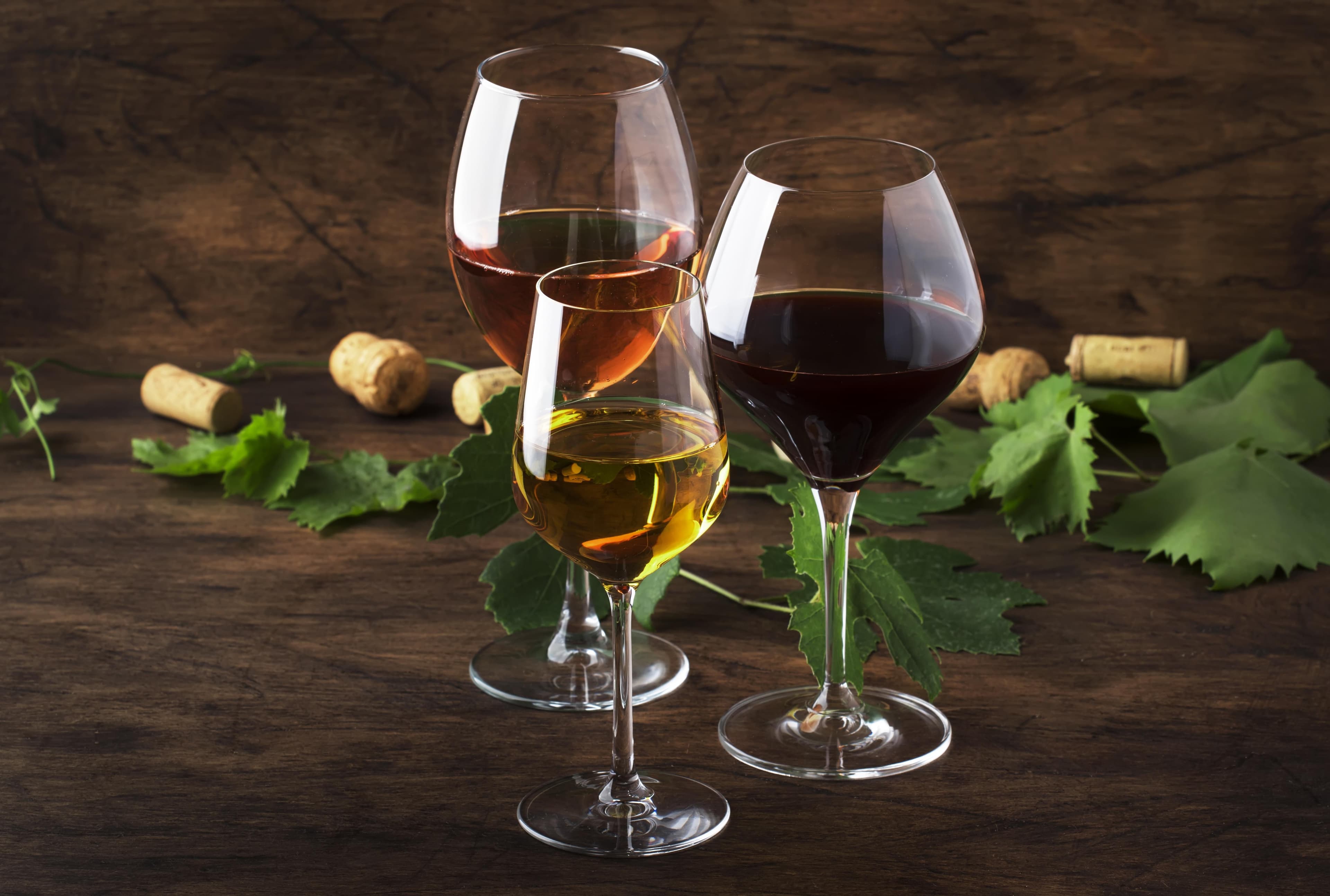 Drei Gläser mit Rot-, Weiß- und Roséwein auf Holz zwischen Weinkorken und Rebenblättern.