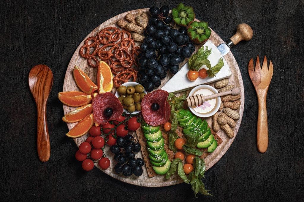 Reichhaltige Snackplatte mit Käse, Salami, Avocado, Weintrauben, Brezeln und Nüssen auf einem Holzbrett.
