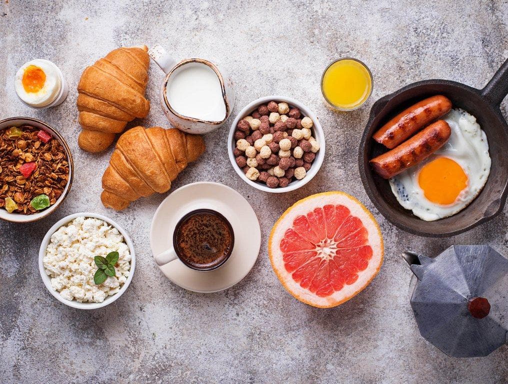 Vielfältiges Frühstück mit Croissants, Spiegelei, Würstchen, Müsli, Kaffee und einer halben Grapefruit in der Draufsicht.