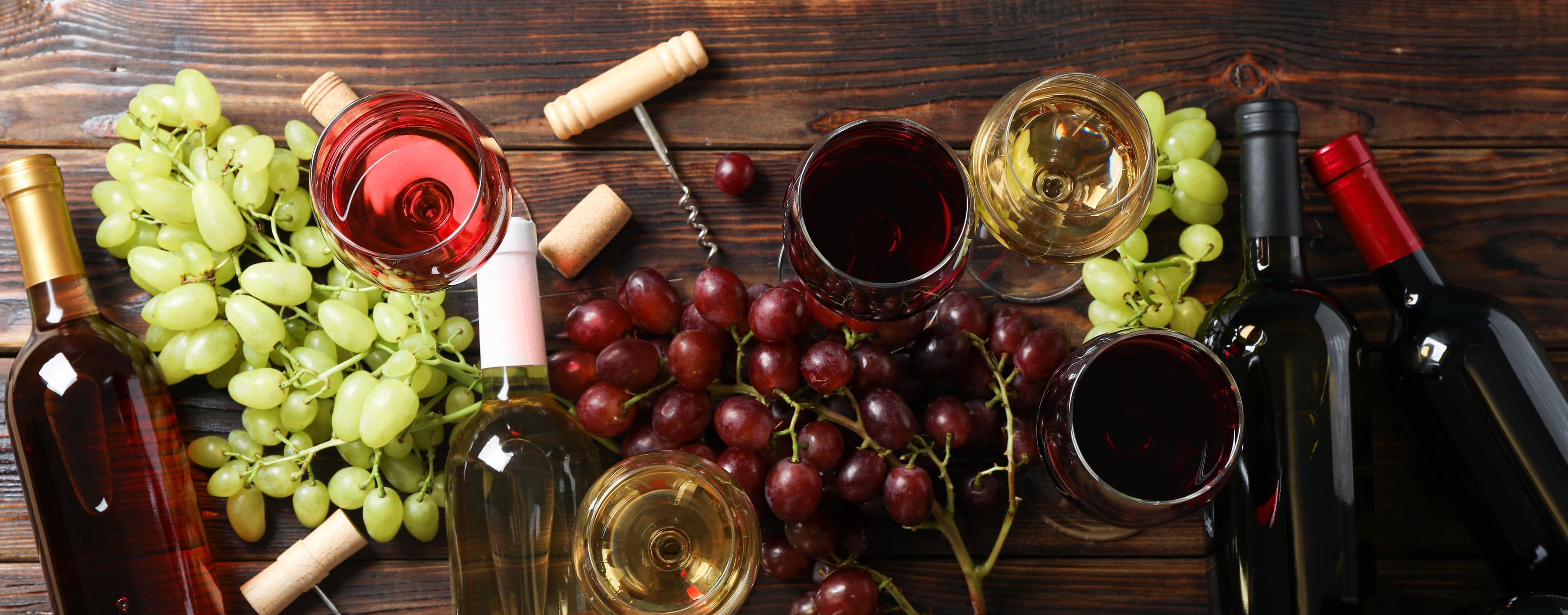 Draufsicht auf verschiedene Weinflaschen, Gläser mit Rot- und Weißwein sowie helle und dunkle Weintrauben auf rustikalem Holz.