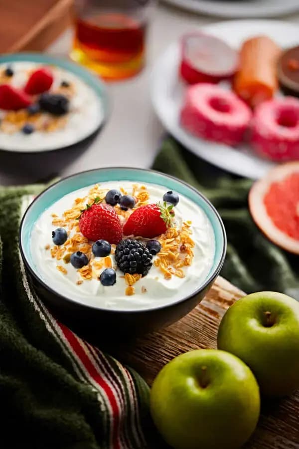 Joghurt-Bowl mit Müsli, Erdbeeren, Blaubeeren und Brombeeren neben grünen Äpfeln auf einem rustikalen Holztisch.