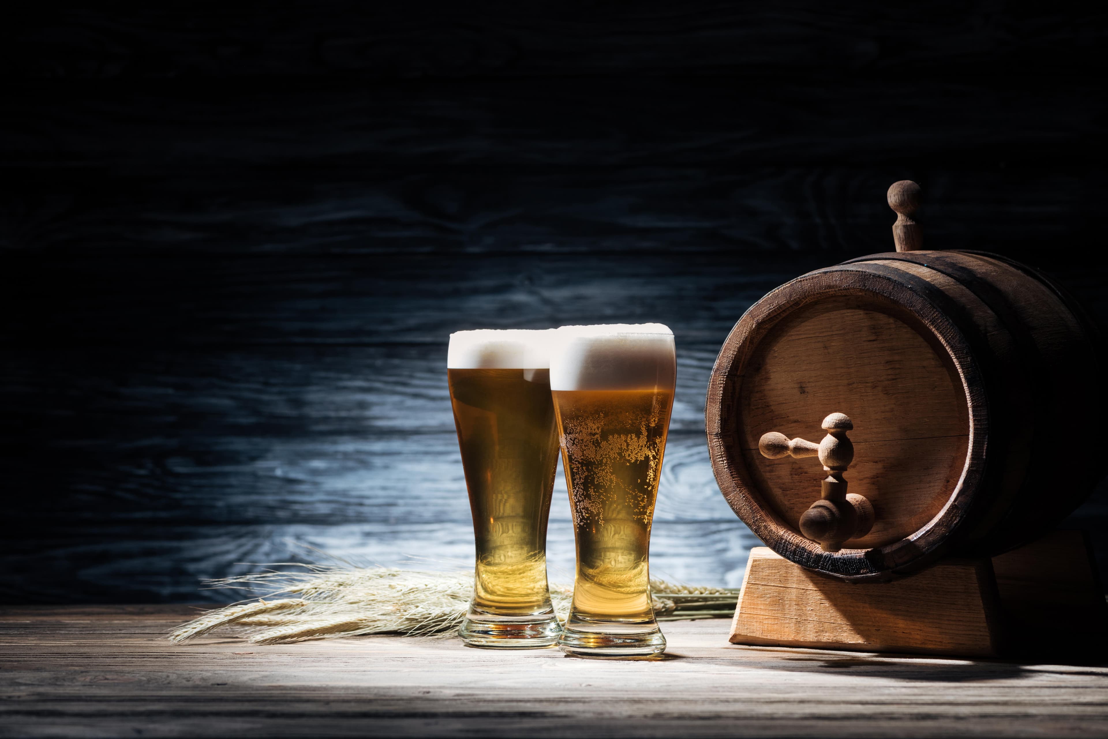 Zwei Gläser frisch gezapftes Bier mit Schaumkrone neben einem kleinen Holzfass und Getreideähren auf rustikalem Holz.