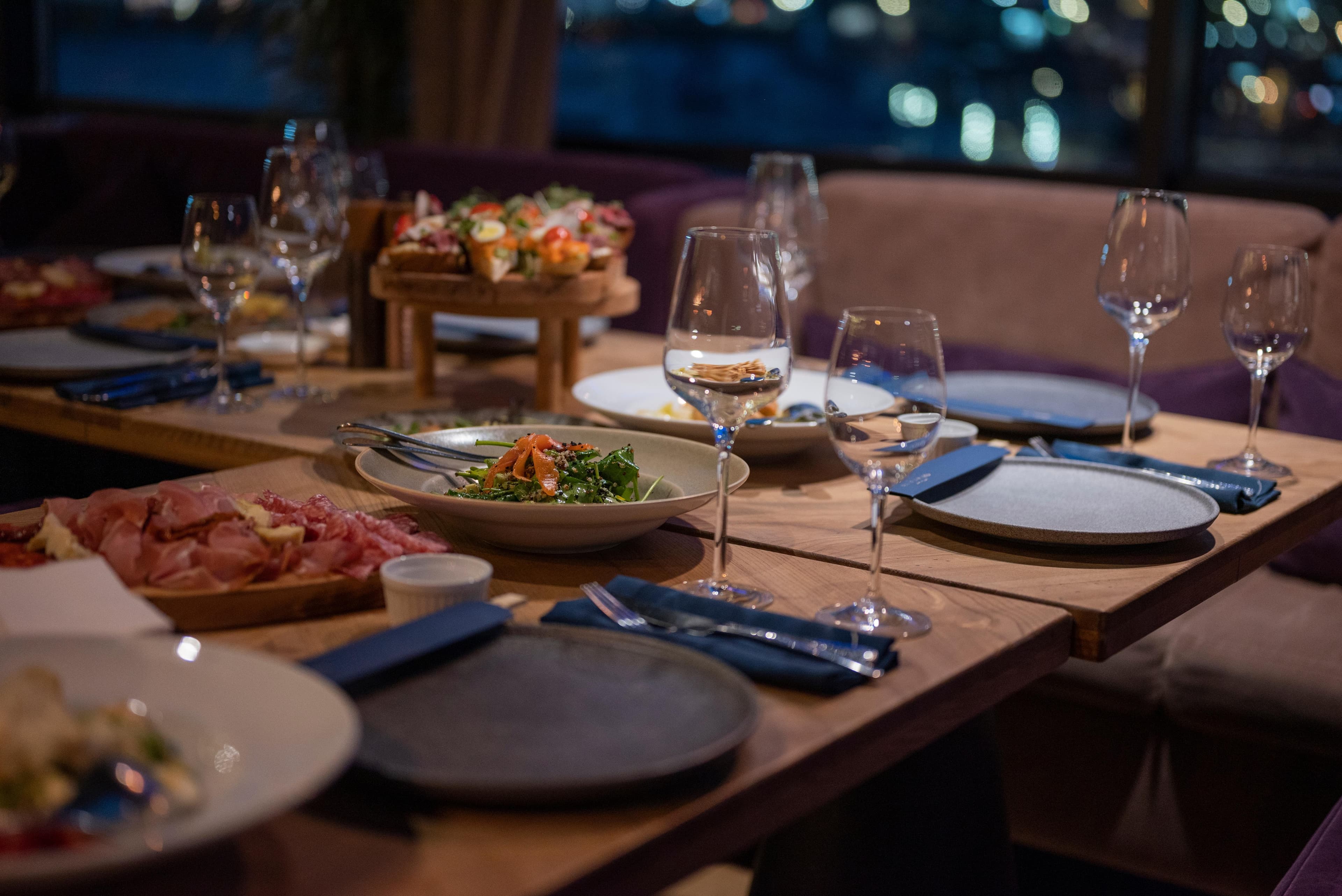 Gedeckter Abendtisch mit Weingläsern, Vorspeisenplatten, Salaten und Aufschnitt in einem Restaurant mit Stadtblick.