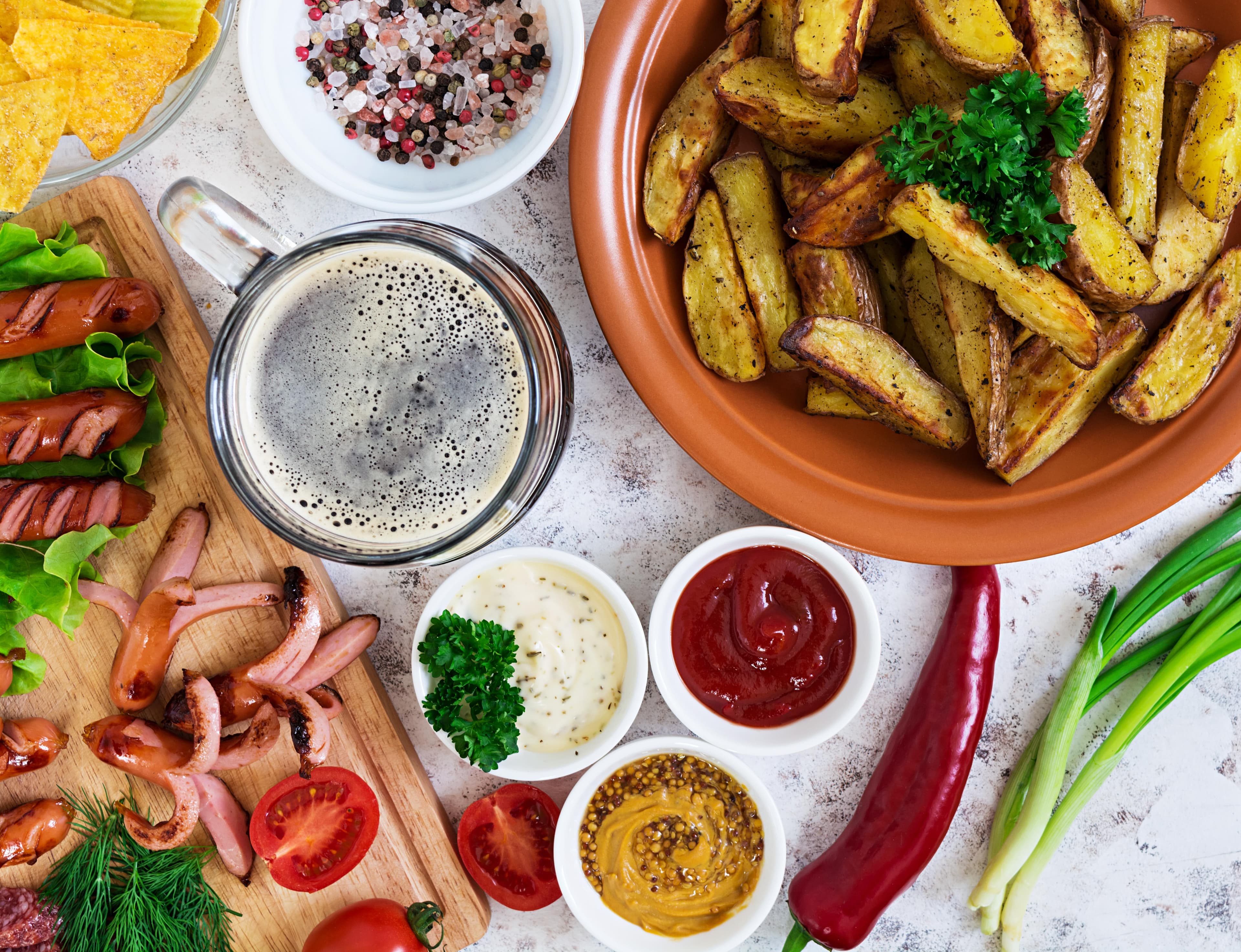 Deftiges Gedeck mit goldbraunen Kartoffelspalten, gegrillten Würstchen, einem Glas dunklem Bier und verschiedenen Dips.