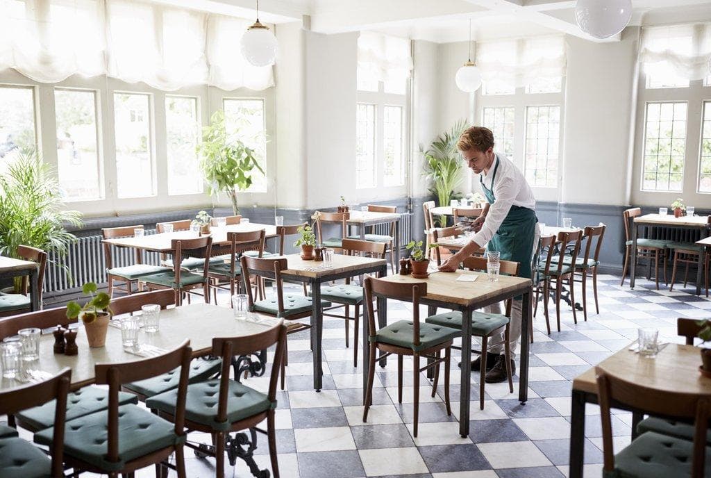 Ein Kellner in grüner Schürze deckt Tische in einem hellen, lichtdurchfluteten Restaurant mit schwarz-weißem Karoboden.