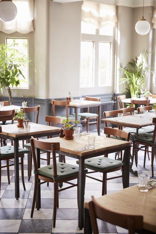 Helles Café mit Holztischen, grünen Polsterstühlen und Schachbrettboden in lichtdurchflutetem Ambiente.