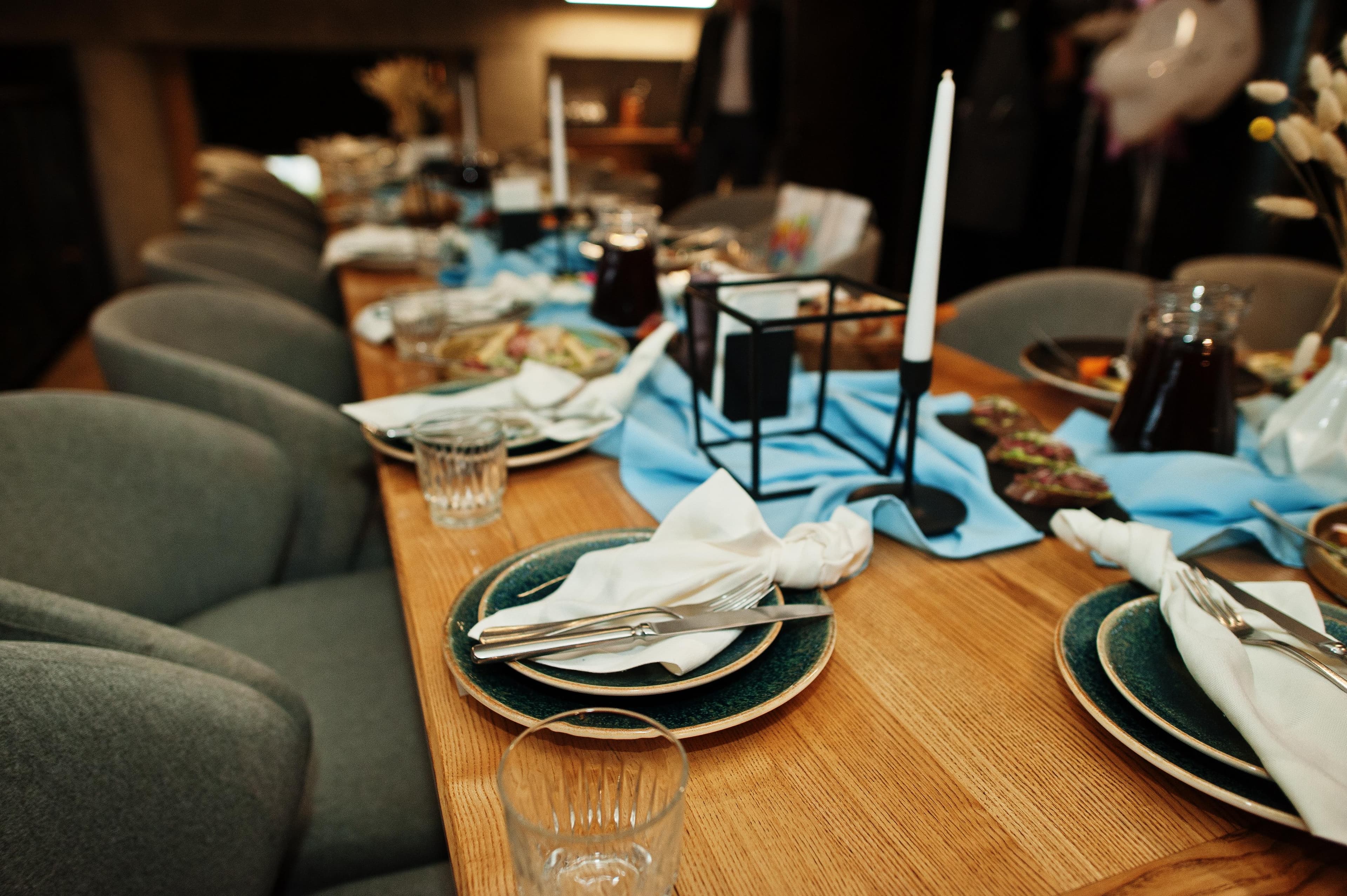 Lange Holztafel mit blauen Läufern, dunklen Tellern, weißen Servietten und Kerzenständern für ein Bankett.