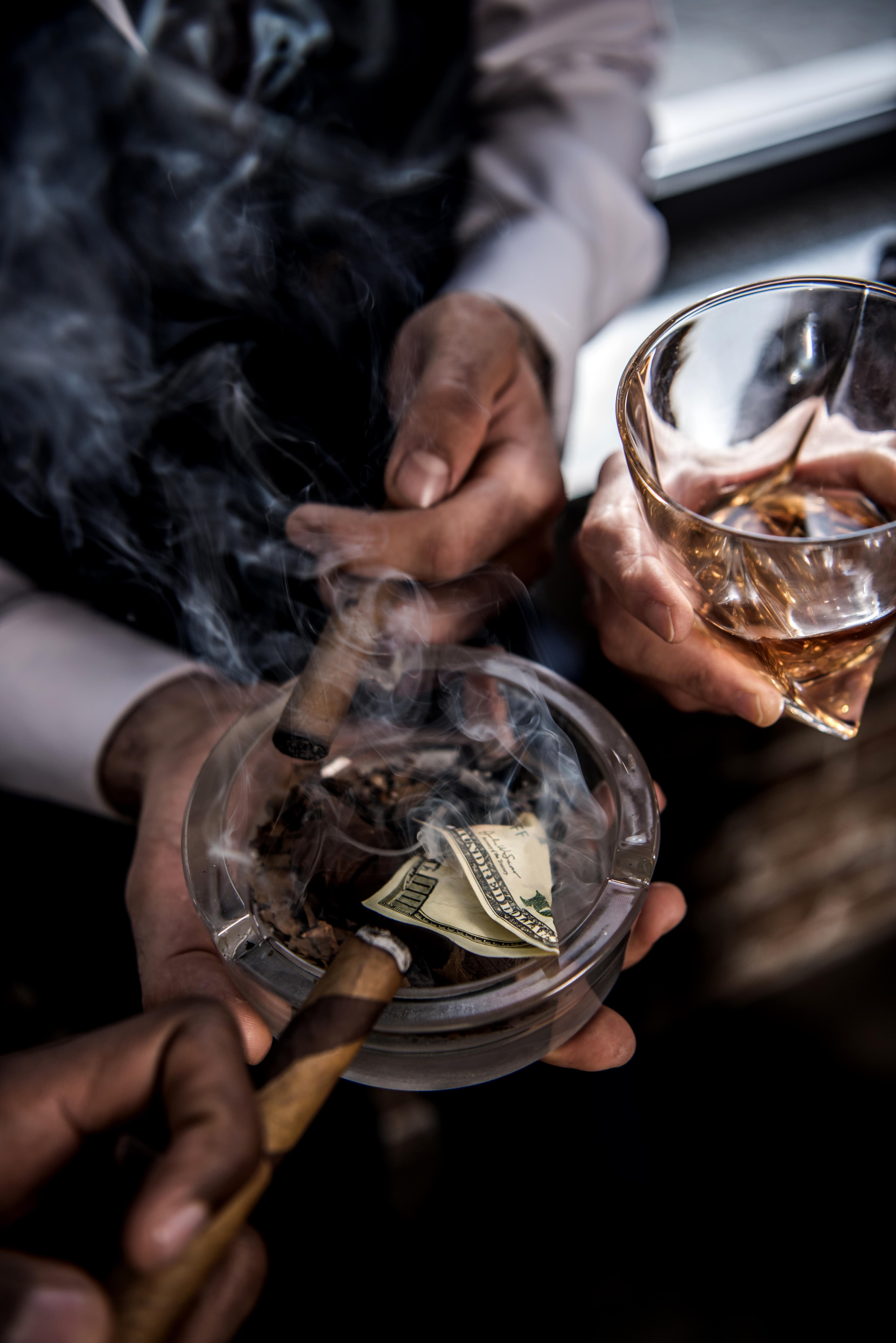 Mehrere Personen halten brennende Zigarren über einen Glasaschenbecher, in dem ein brennender 100-Dollar-Schein liegt.
