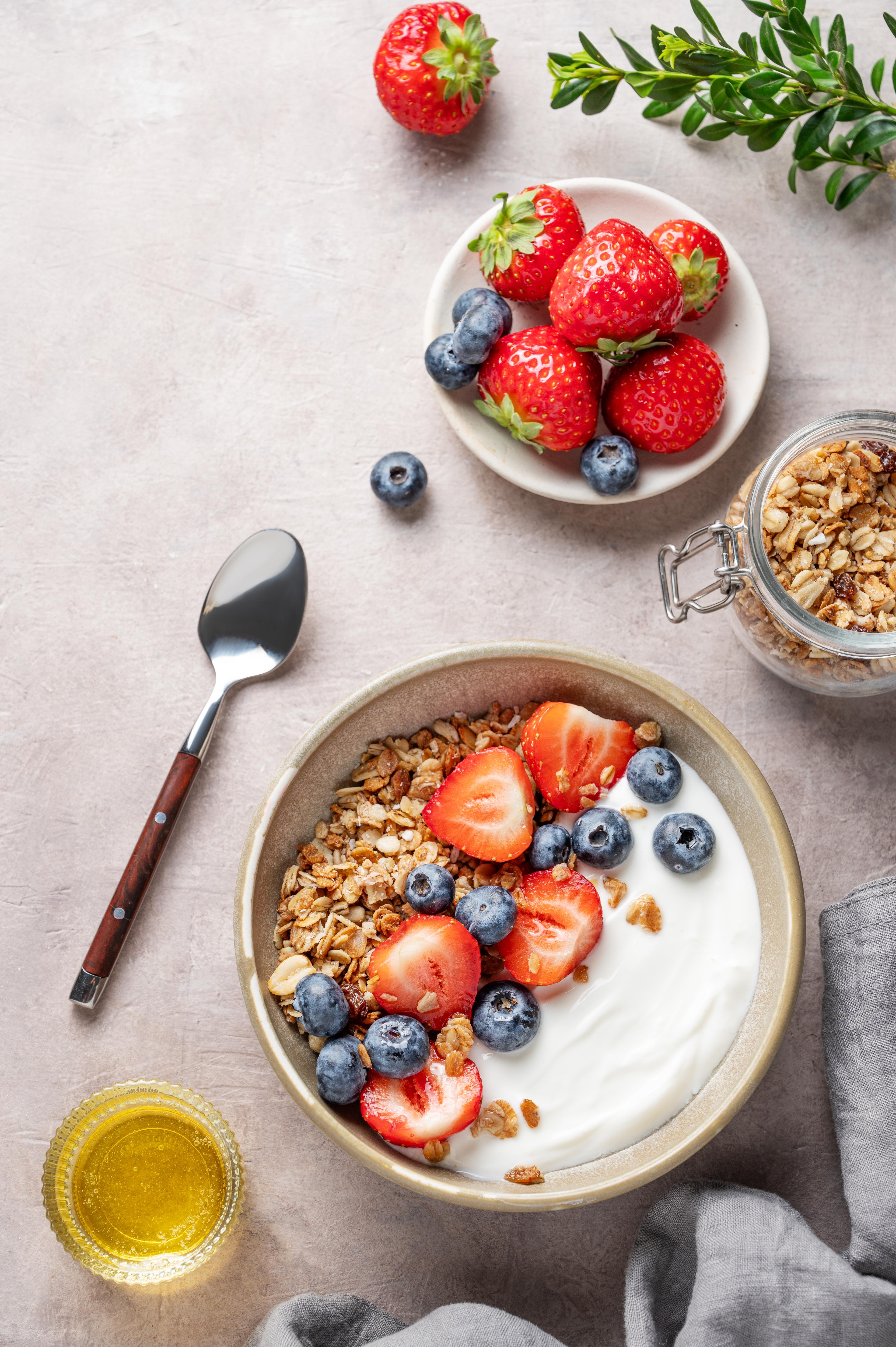 Müsli-Bowl mit Joghurt, frischen Erdbeeren und Blaubeeren, serviert mit Honig und Knuspermüsli in der Draufsicht.