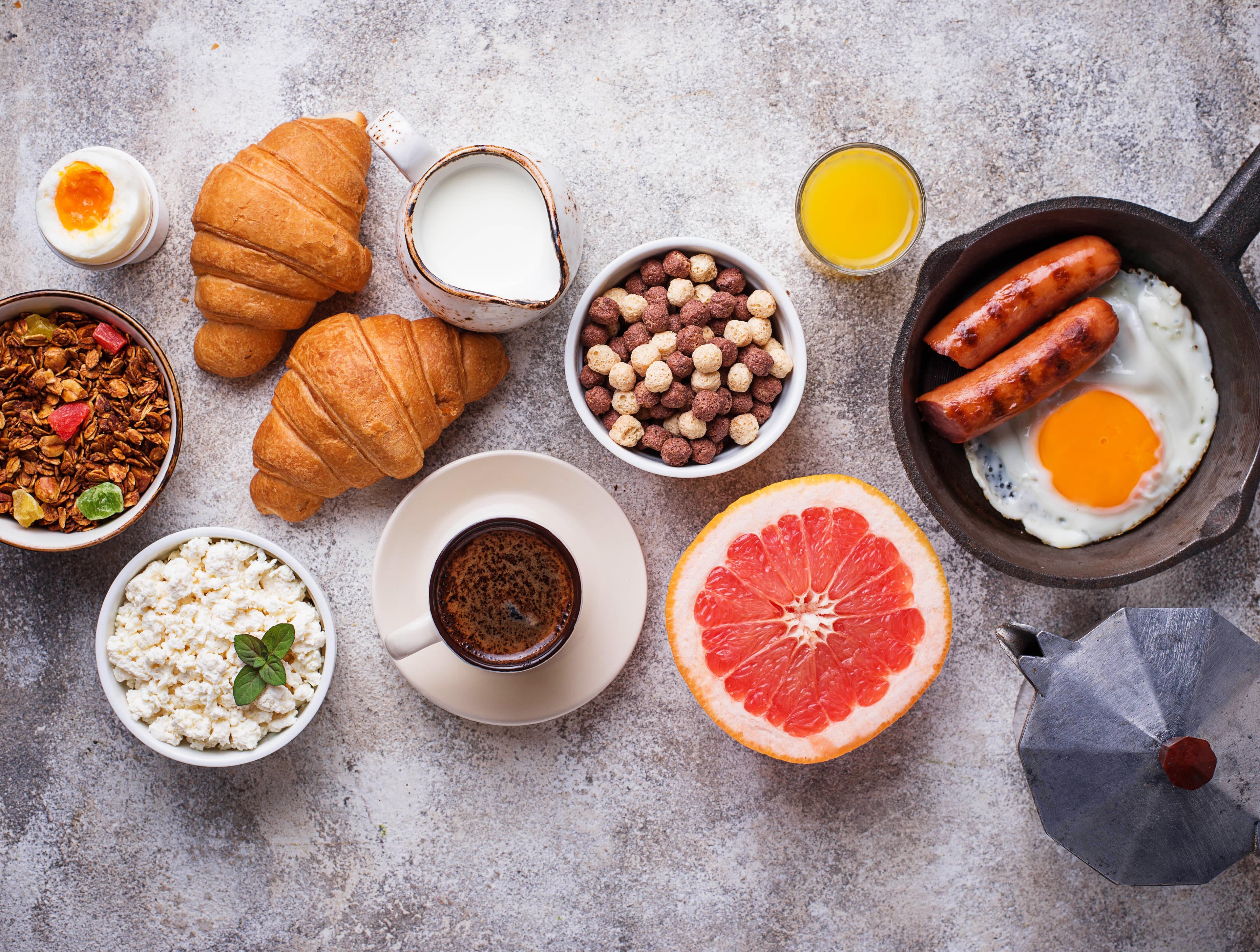 Vielfältiges Frühstück mit Croissants, Spiegelei, Würstchen, Müsli, Kaffee und einer halben Grapefruit in der Draufsicht.