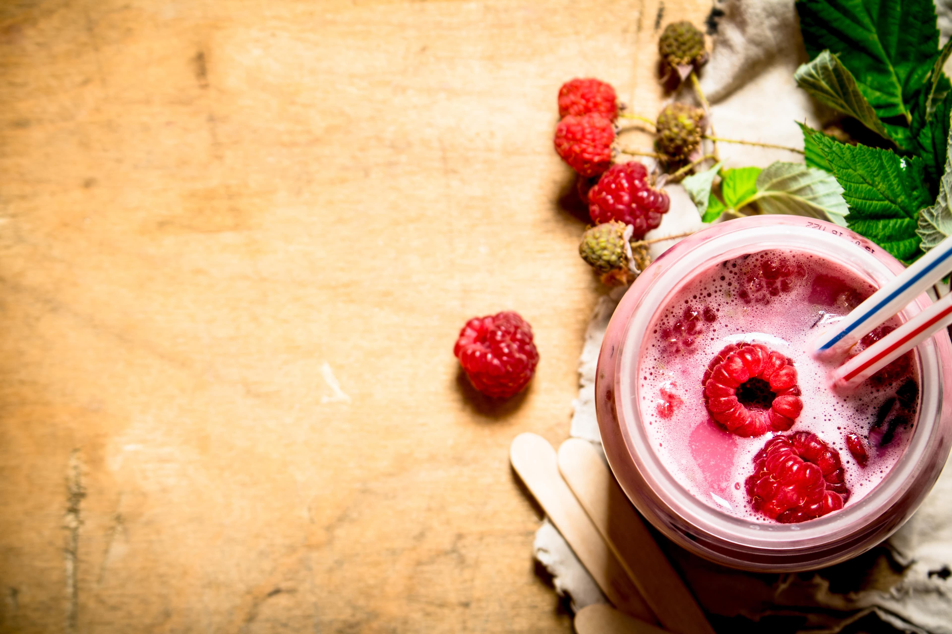 Ein Glas pinker Himbeer-Smoothie mit frischen Beeren und Strohhalmen auf einem rustikalen Holzhintergrund.