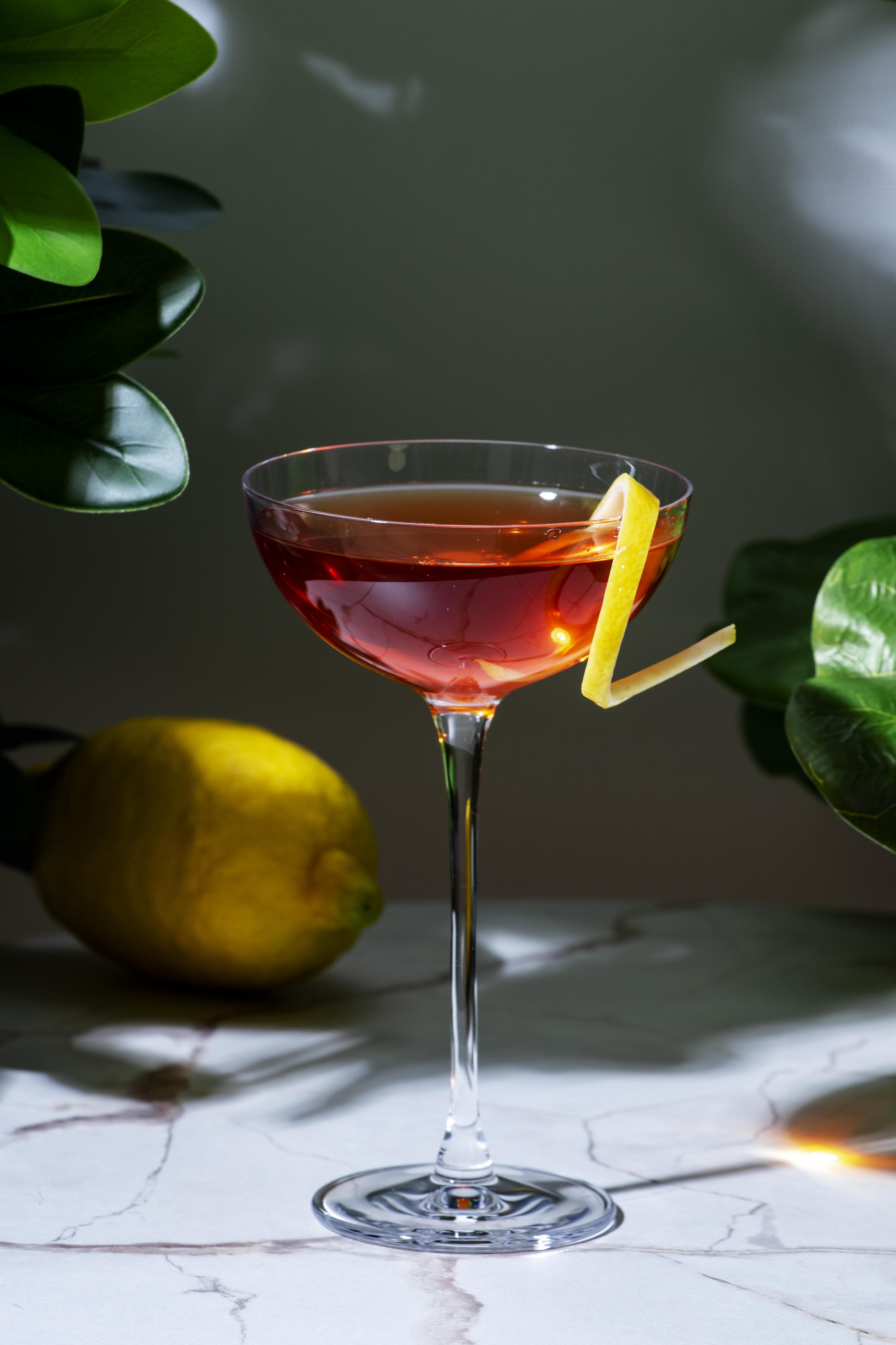 Ein bernsteinfarbener Cocktail in einer flachen Sektschale, garniert mit einer langen Zitronenzeste vor einer Marmoroberfläche.