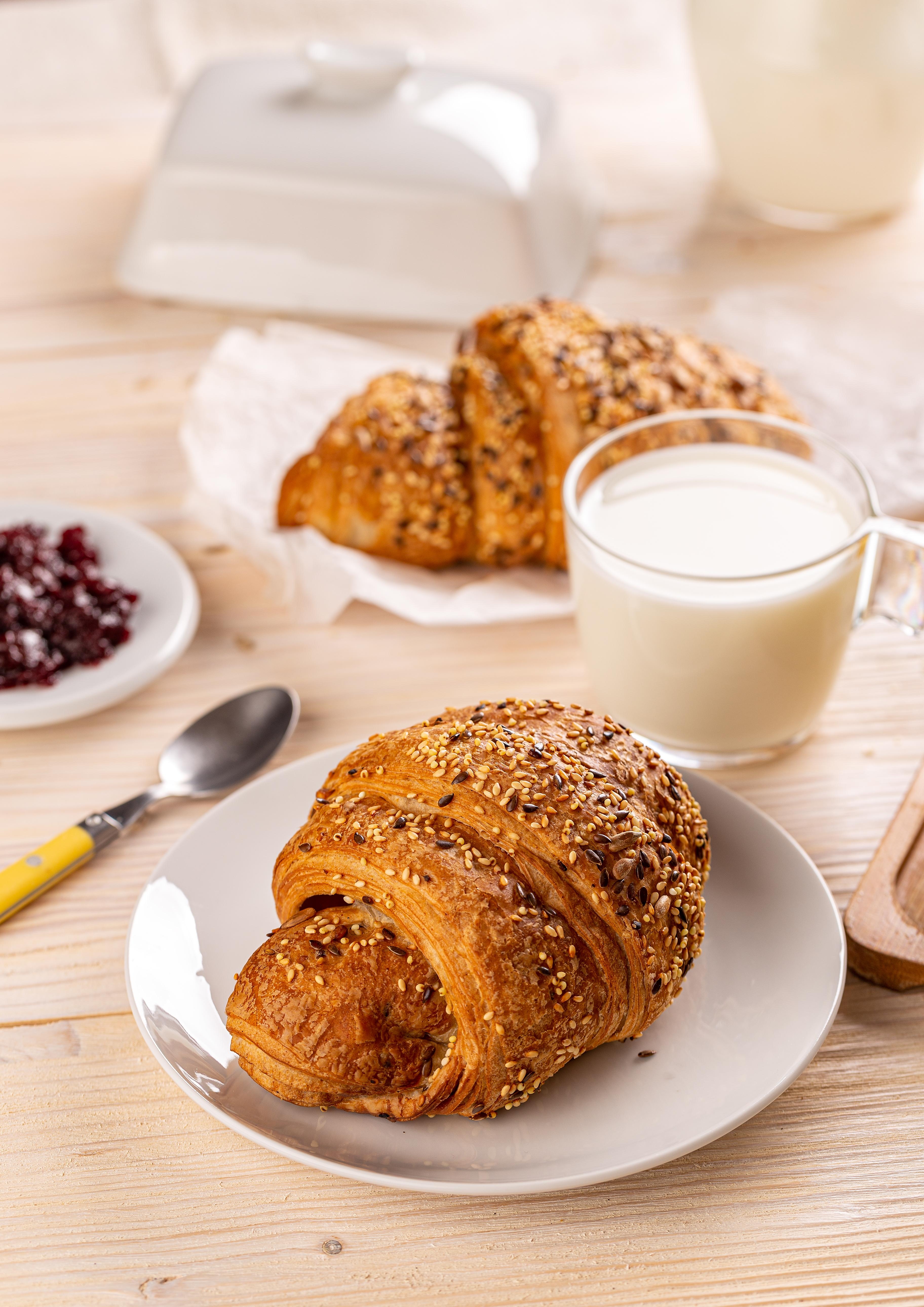 Frisches Laugen-Croissant mit Saaten auf einem Teller, serviert mit einem Glas Milch und Beerenkonfitüre.