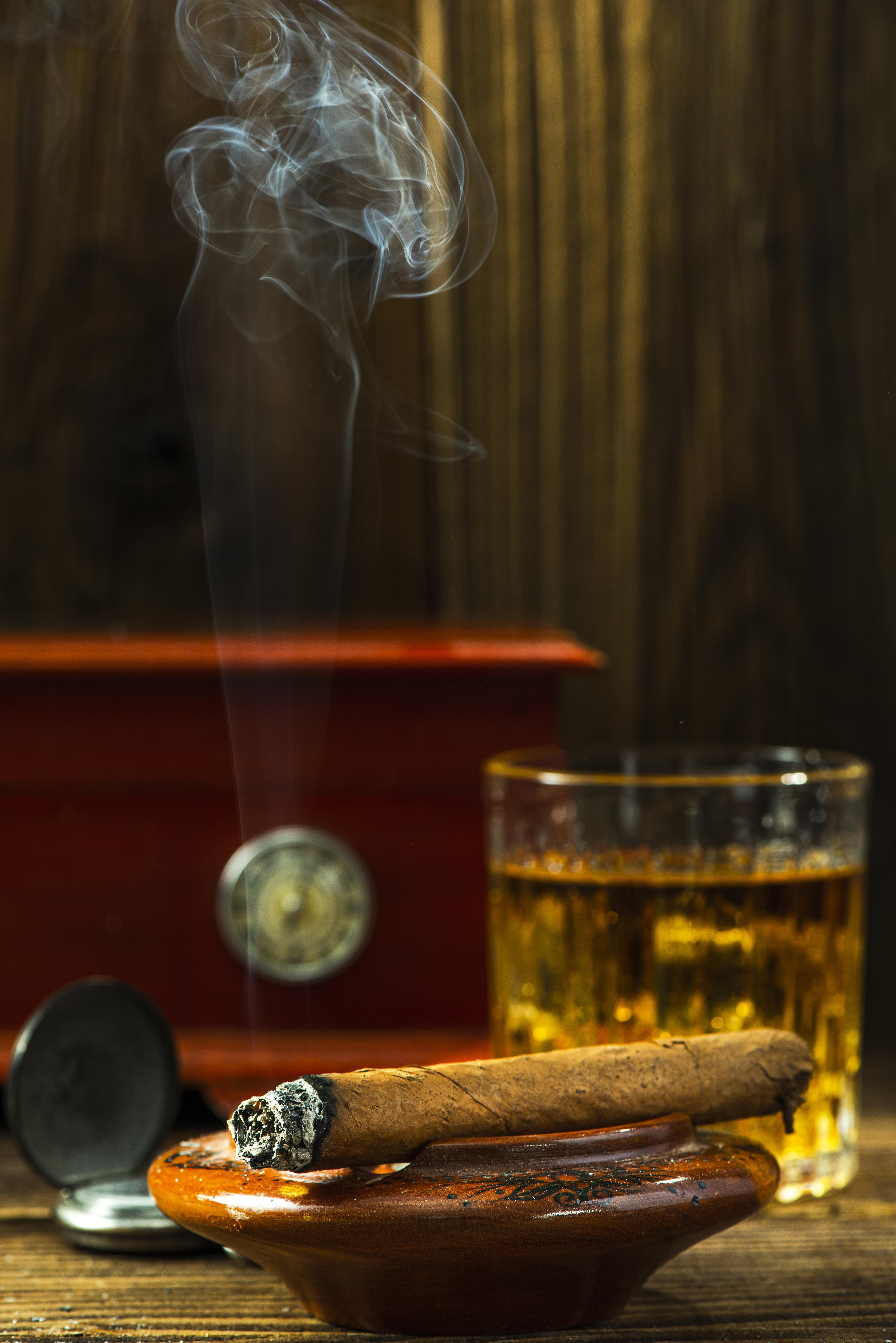 Eine brennende Zigarre liegt in einem Aschenbecher; Rauch steigt vor einem Glas Whiskey und einem Humidor auf.