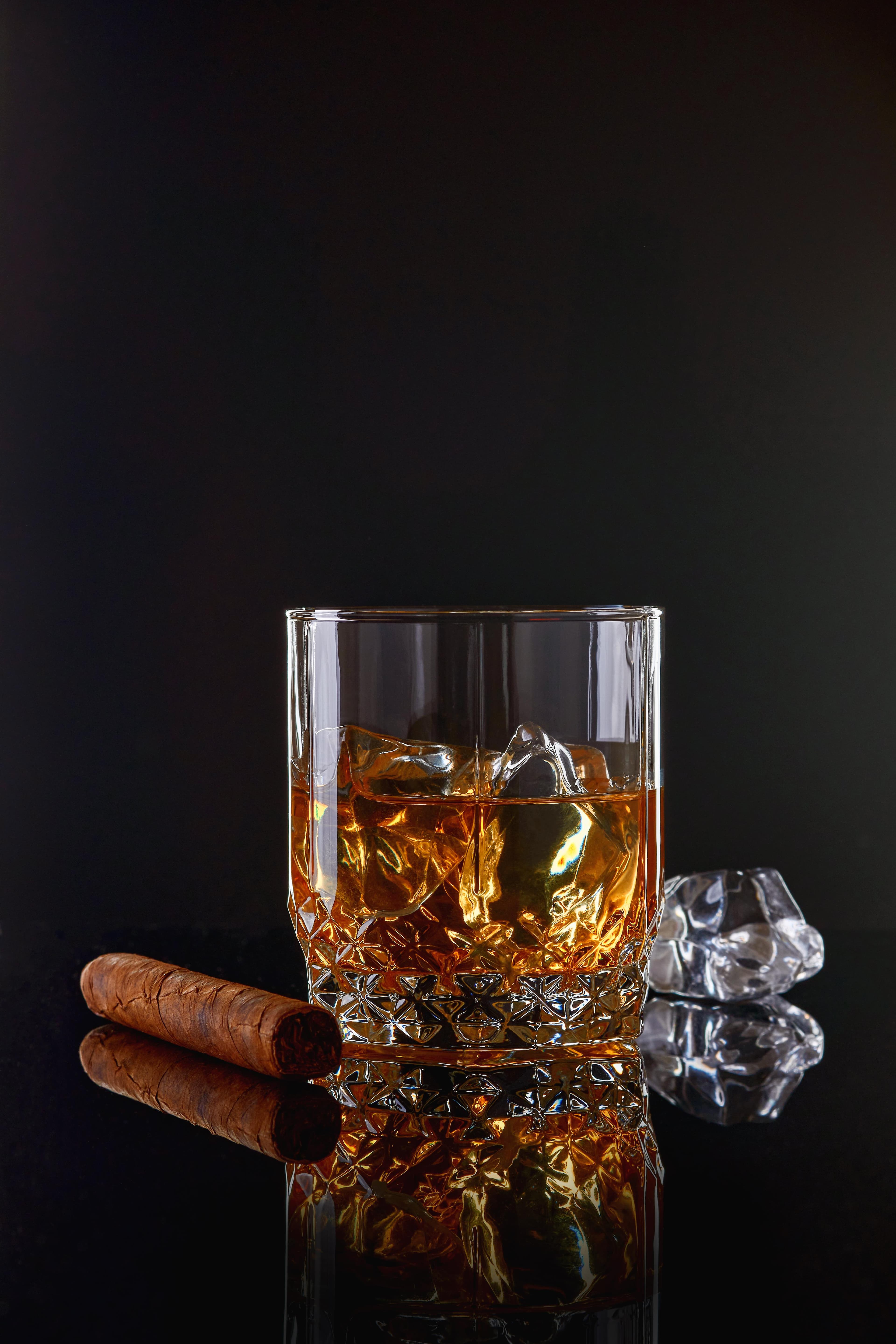 Ein Kristallglas mit Whiskey auf Eis neben einer unangezündeten Zigarre, die sich auf einer dunklen Oberfläche spiegeln.